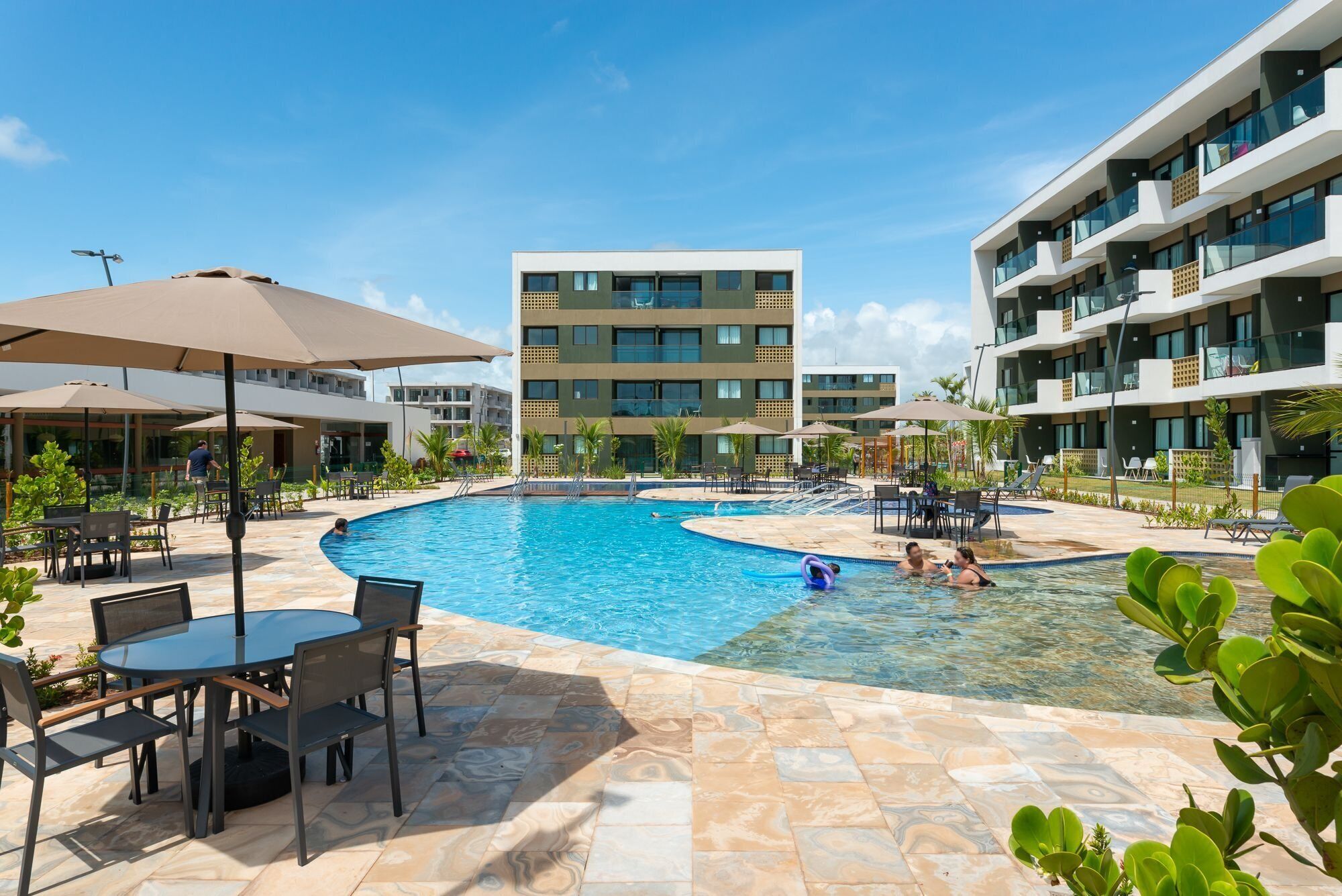 Zona de piscina con sillas de descanso y sombrillas, rodeada de edificios de varios pisos con balcones.
