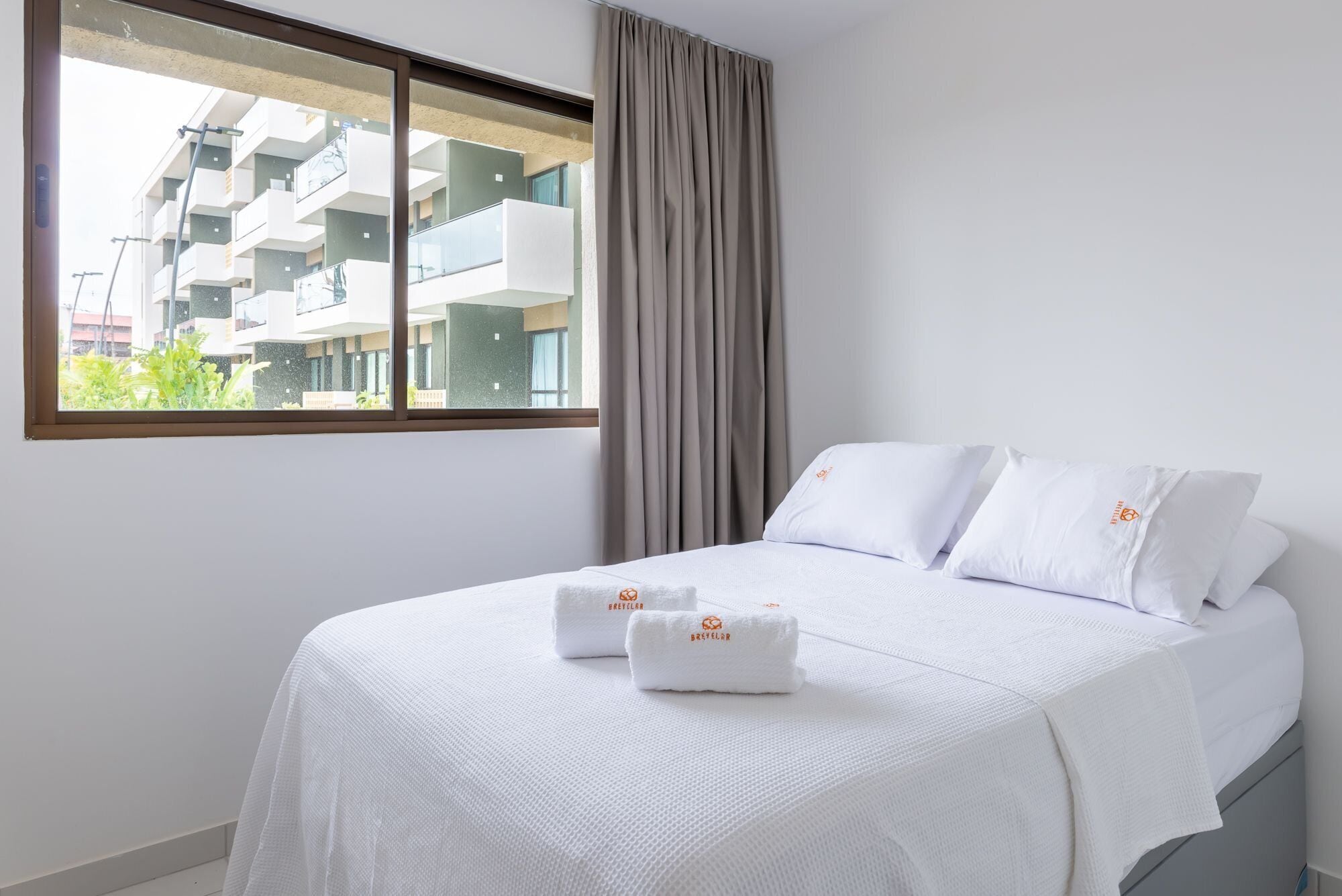 Una habitación de hotel con una cama, ropa de cama blanca, una ventana con vistas a un edificio y una cortina.