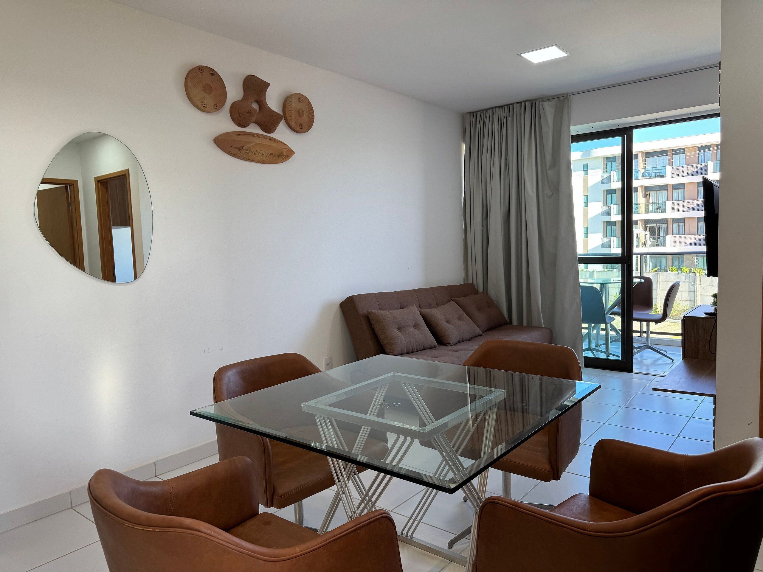 A modern living room with a glass-top table and leather chairs, a sofa with cushions, a round mirror, and a wall-mounted decorative piece.