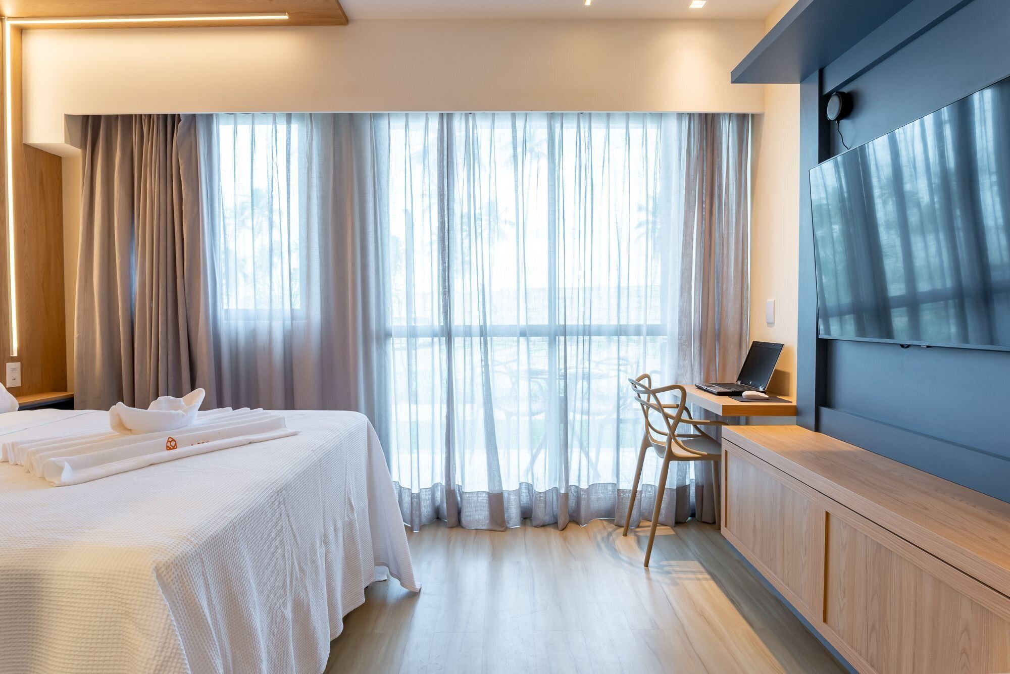 Una habitación de hotel con cama, escritorio y computadora portátil, y un ventanal con cortinas.