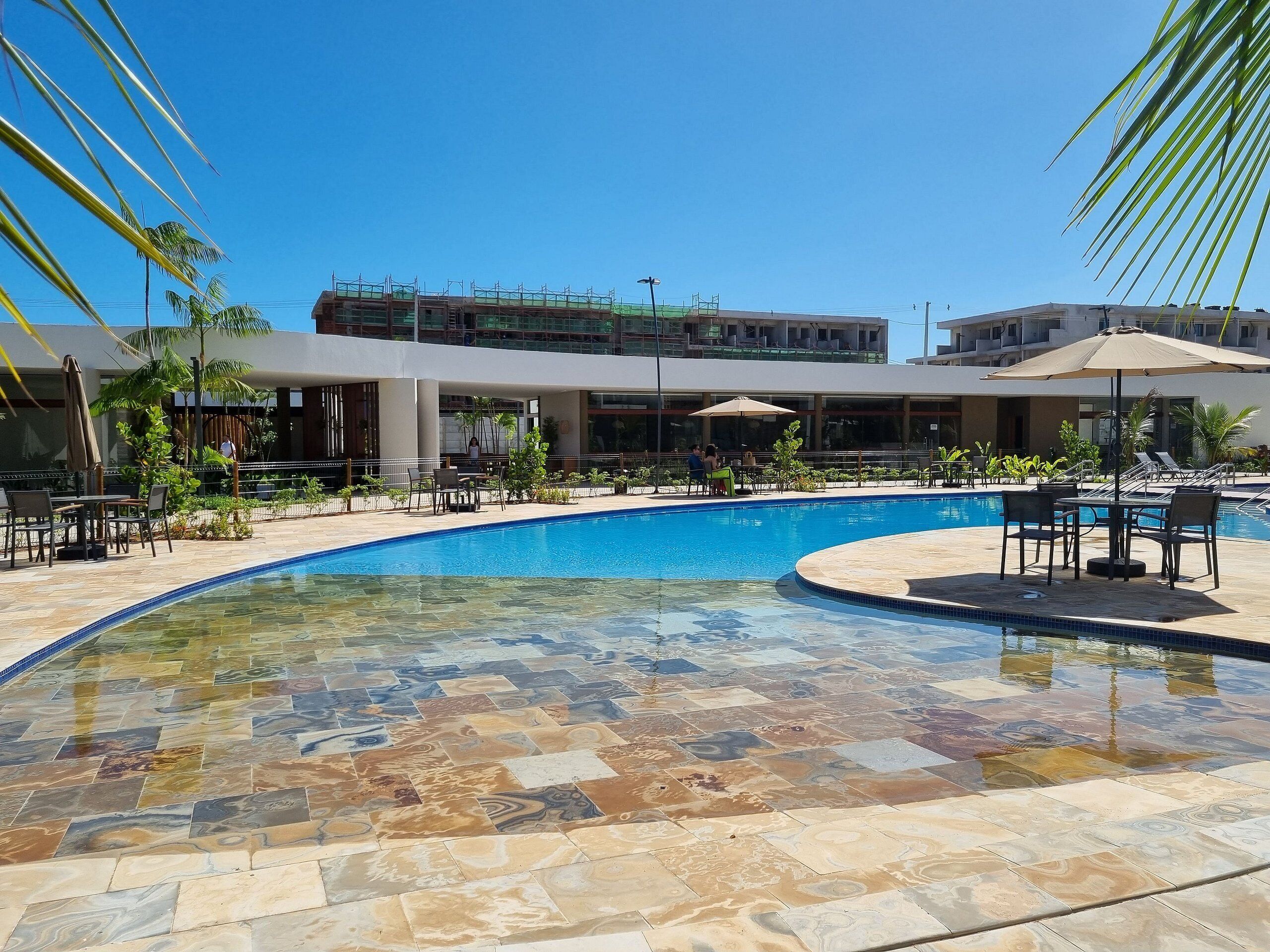 Piscina com deck ladrilhado, cercada por cadeiras de praia e guarda-sóis, com um edifício ao fundo.