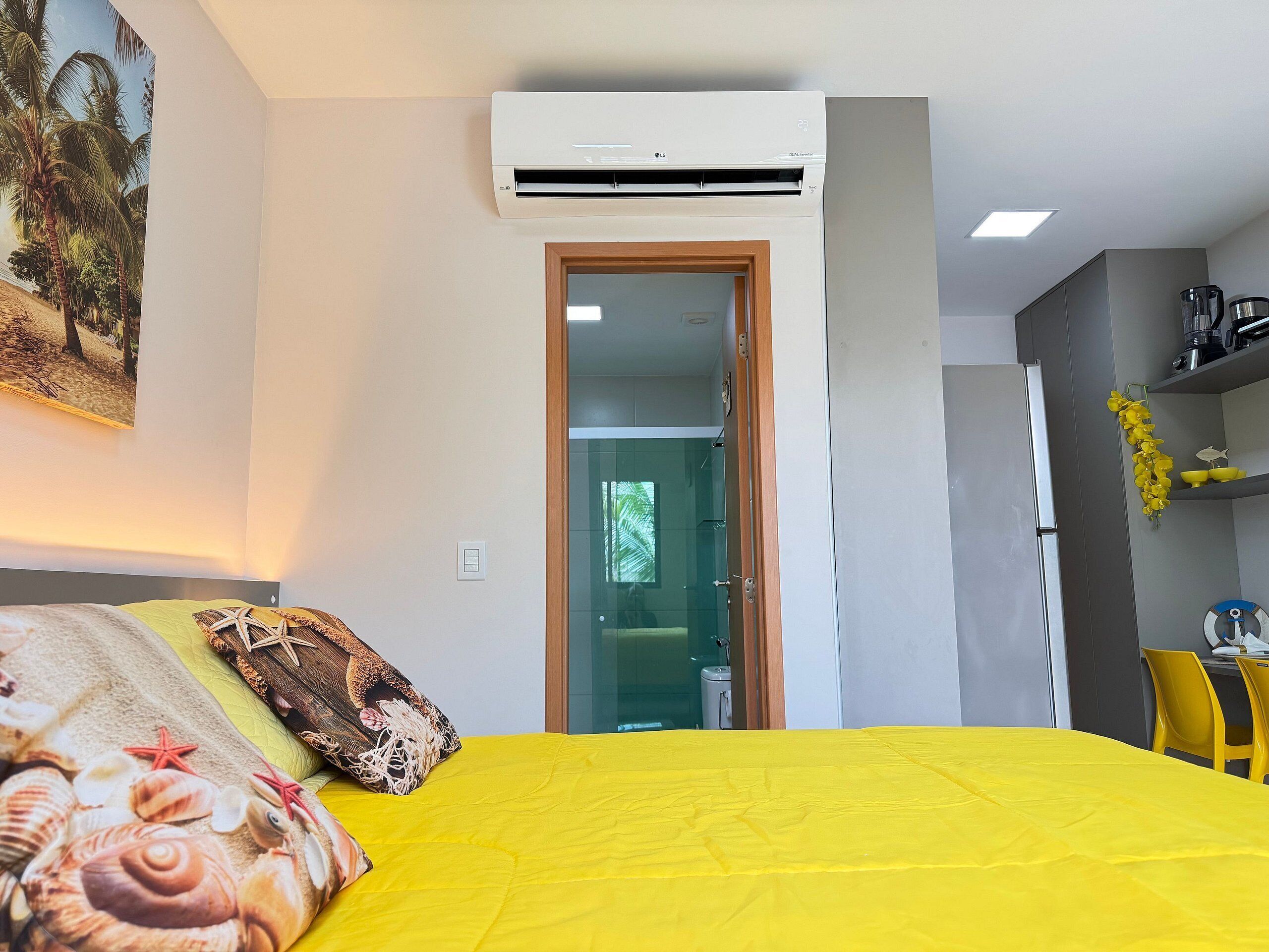 Una habitación con cama, aire acondicionado, una puerta que da a otra habitación y una zona de cocina con refrigerador y lavabo.