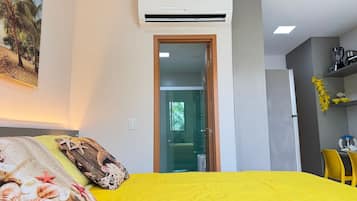 Una habitación con cama, aire acondicionado, una puerta que da a otra habitación y una zona de cocina con refrigerador y lavabo.