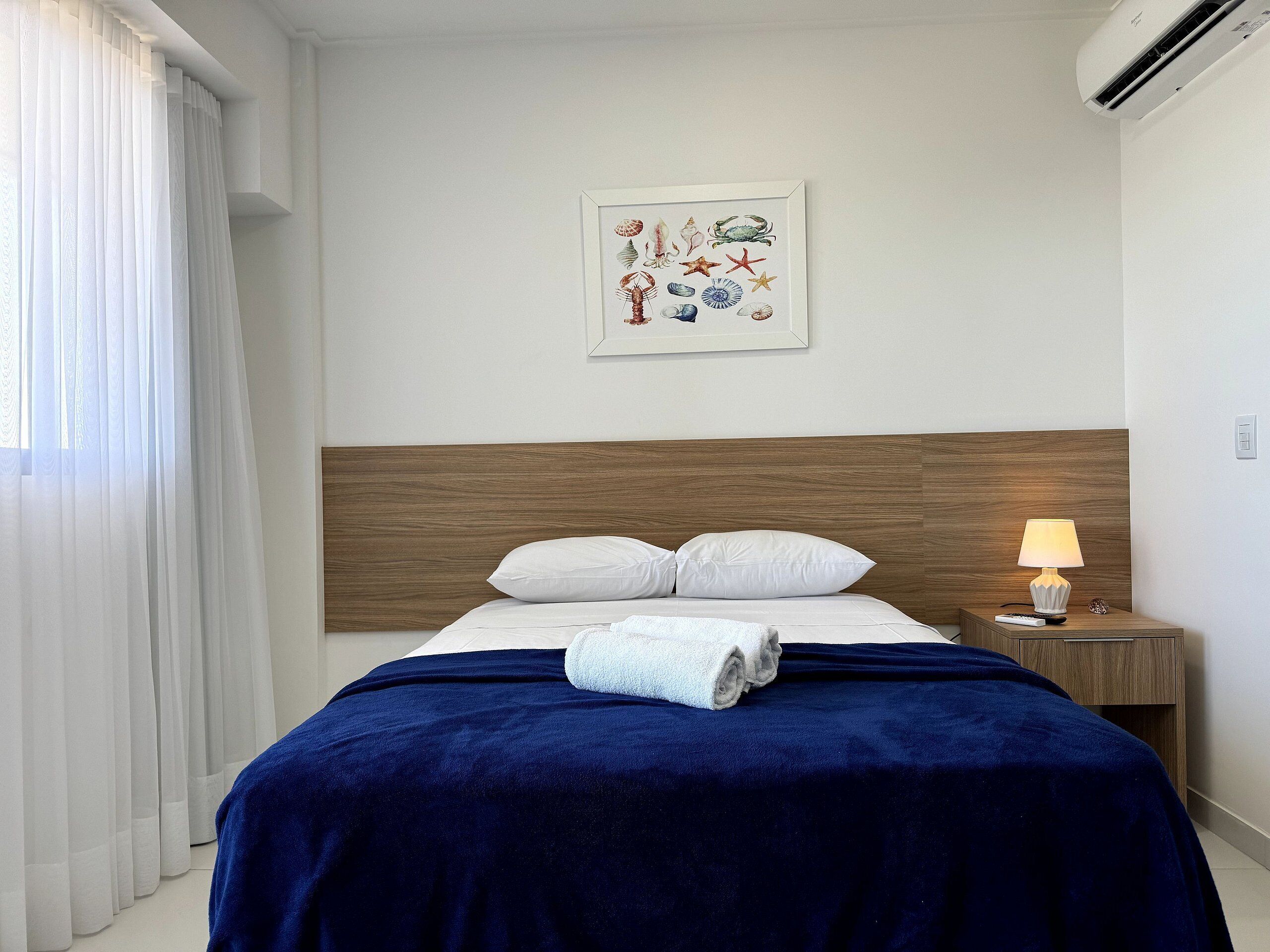 A hotel room with a bed, a nightstand with a lamp, a framed picture on the wall, and a window with curtains.