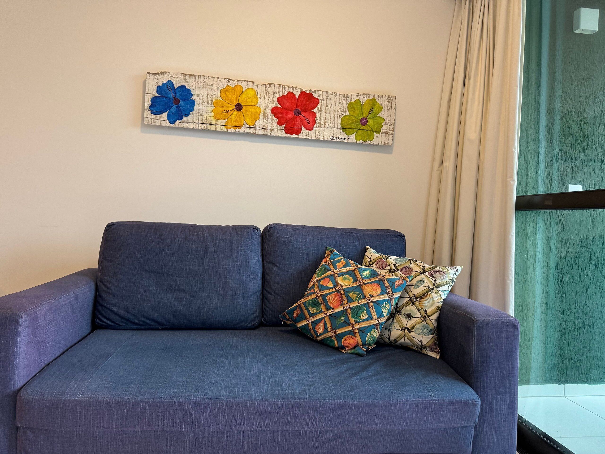 Ein blaues Sofa mit gemusterten Kissen, ein blumiges Wandbild und ein Fenster mit Vorhängen.