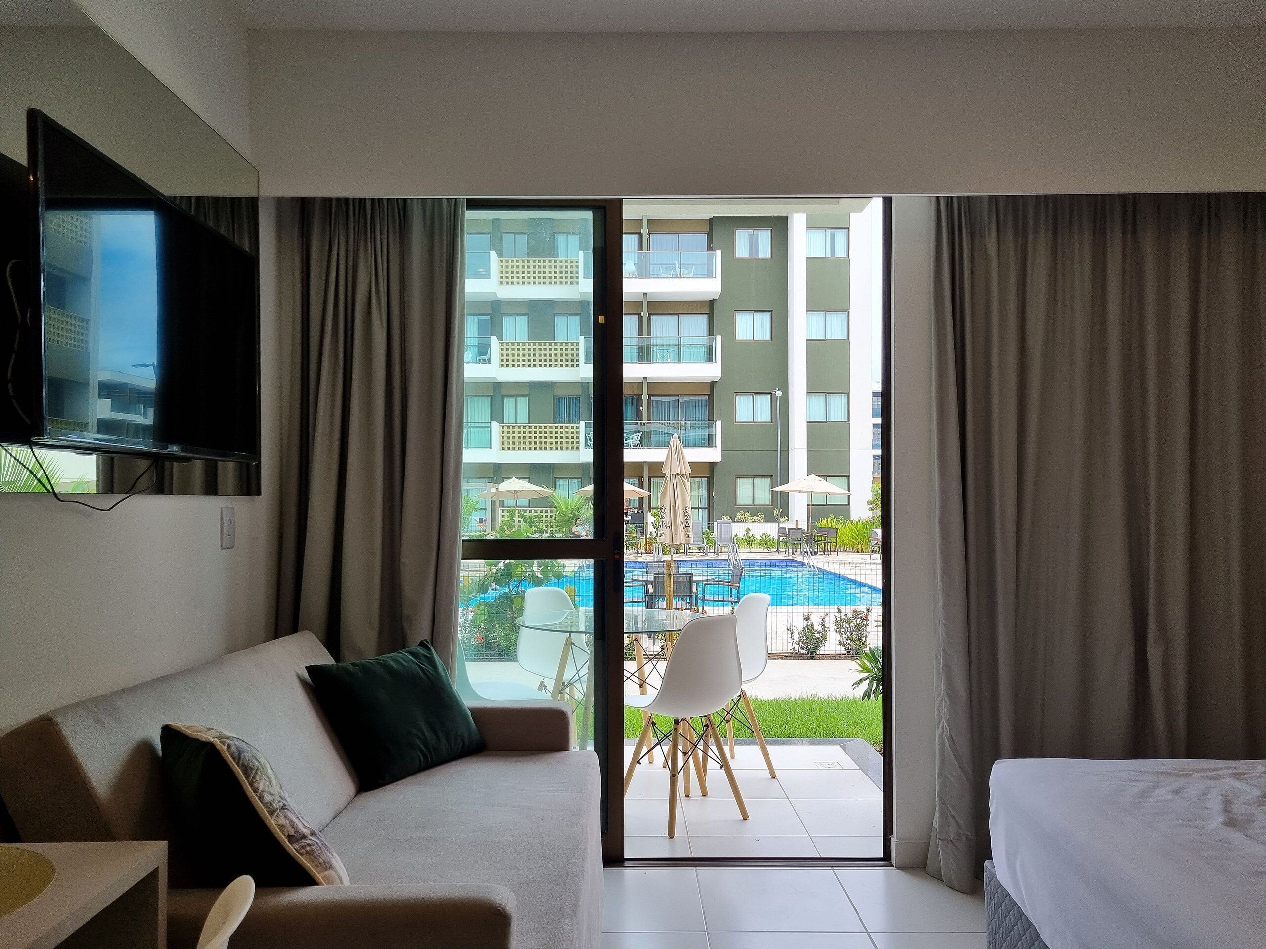 A hotel room with a sofa, a bed, a TV mounted on the wall, and a view of a swimming pool and building.