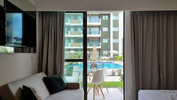A hotel room with a sofa, a bed, a TV mounted on the wall, and a view of a swimming pool and building.