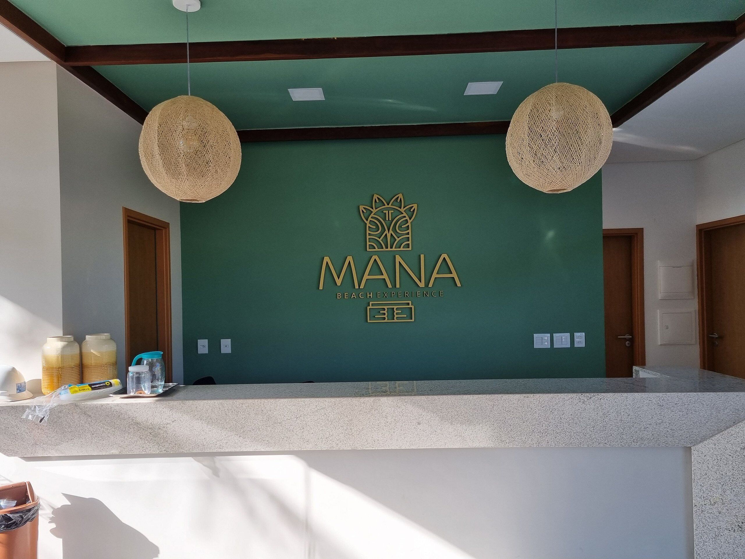 Un mostrador de recepción con una pared verde, dos lámparas tejidas y el logo de 'MANA Beach Experience'.
