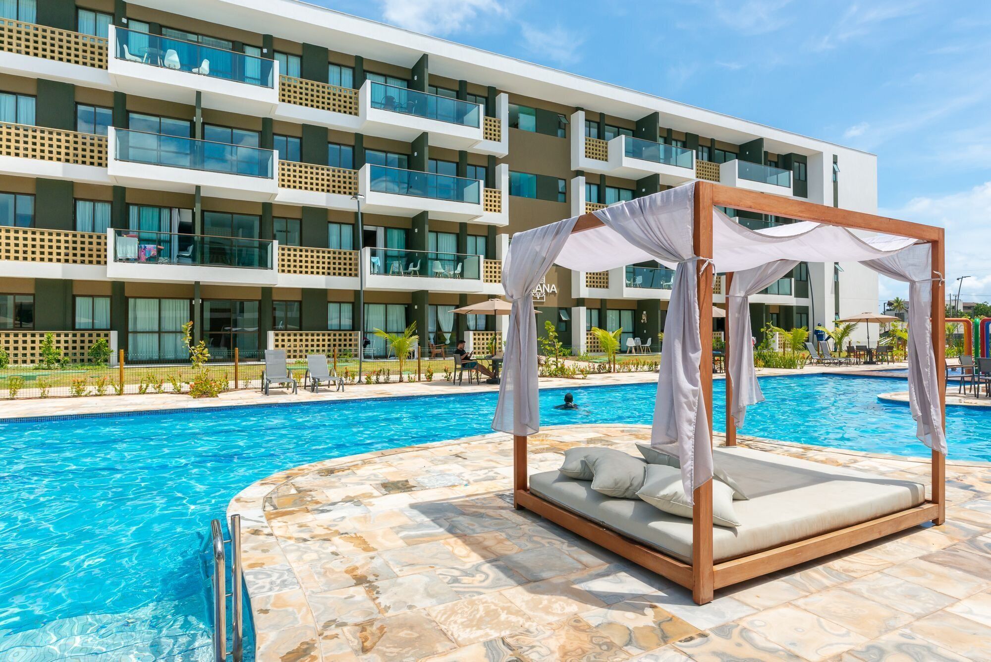 A hotel pool area with a canopy bed, clear blue pool water, and a modern building with balconies in the background.