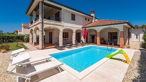 Una casa de dos pisos con piscina, mobiliario de exterior y un paraguas rojo.