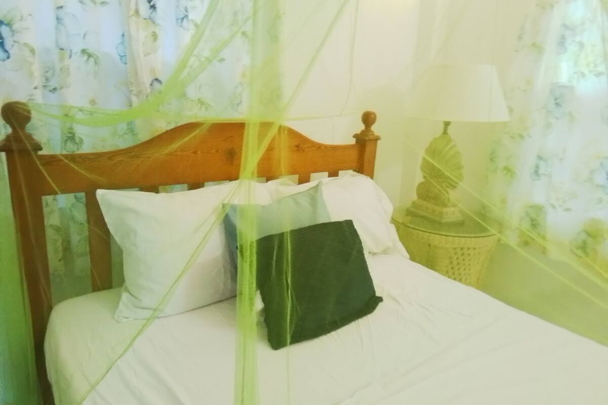 Een bed met een houten hoofdbord, wit beddengoed en een groen kussen. Er staat een lamp op een nachtkastje en een decoratief object aan de muur.