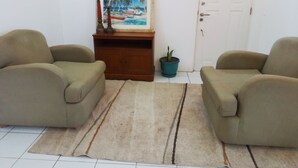 Une pièce avec deux fauteuils beiges, un meuble en bois, un tableau représentant une scène de plage, une plante en pot et une porte blanche avec une fenêtre.