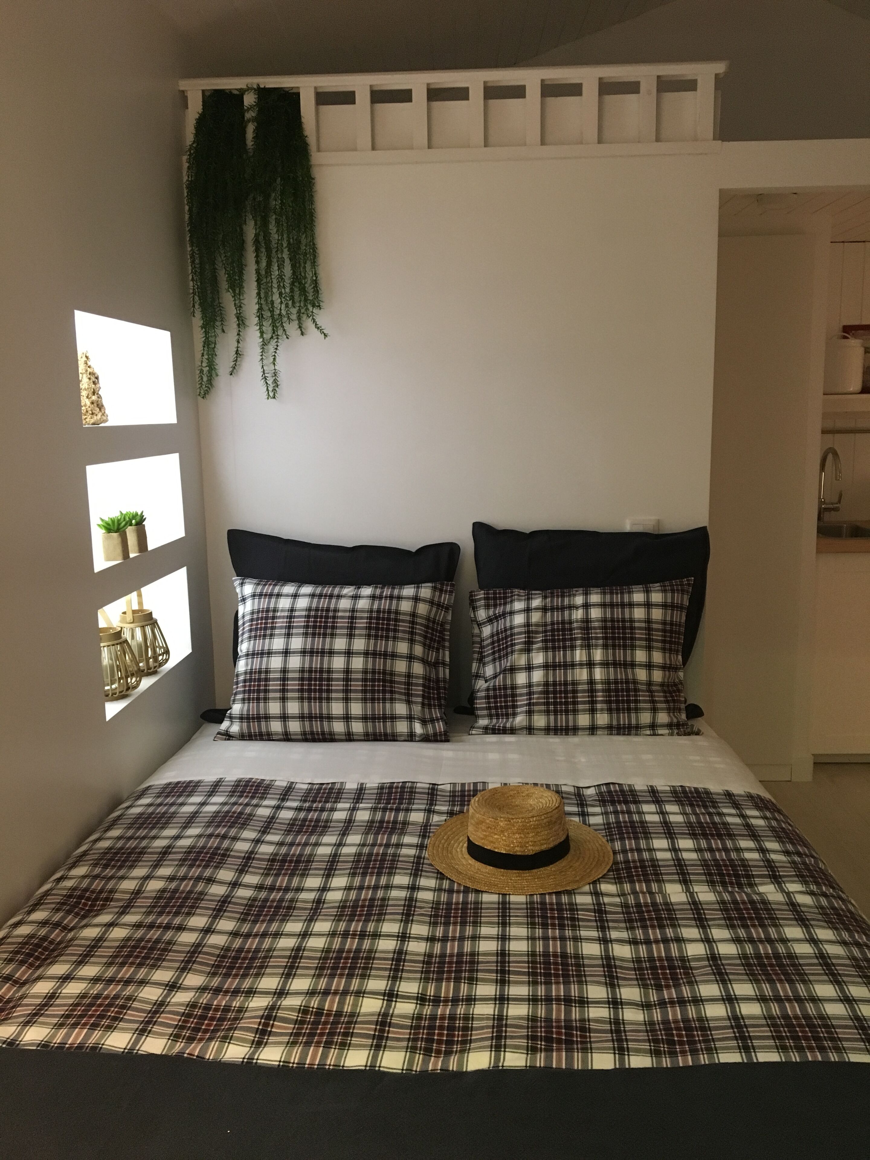 Una cama con ropa de cuadros, dos almohadas, un sombrero y una ventana con plantas.