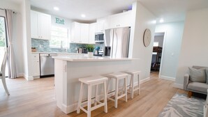 Una cocina moderna con armarios blancos, una isla central con taburetes y una zona de comedor con sofá.