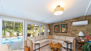 A living room with a large sectional sofa, a round coffee table, and a stone wall. There are two windows and an air conditioning unit on the wall. - Sparkling pool and rejuvenating spa! (Saint Augustine)