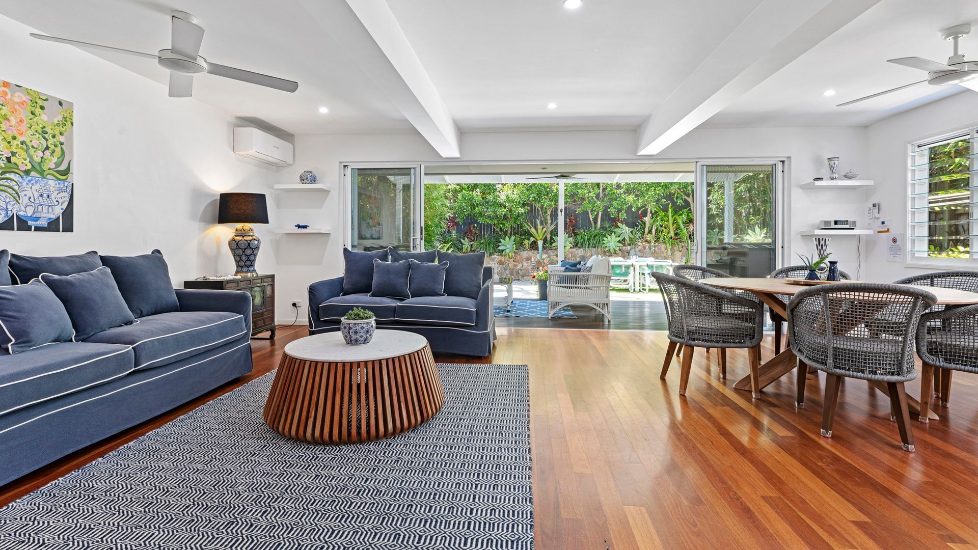 A modern living room with a blue sofa, a round wooden coffee table, and a dining area with wicker chairs and a wooden table.