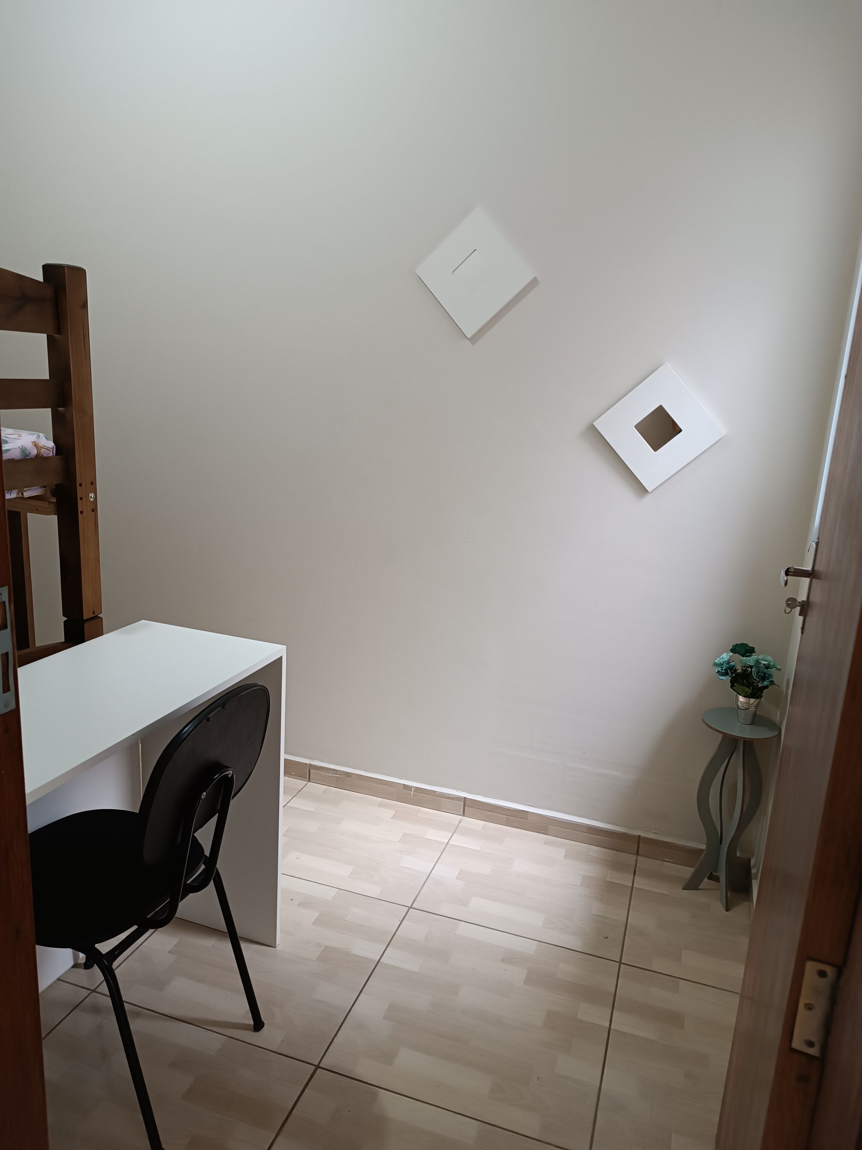 Et lille, rent værelse med et hvidt skrivebord, en sort stol og et lille bord med en plante.