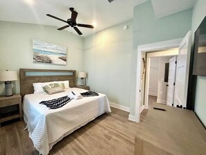 A bedroom with a bed, nightstands, a ceiling fan, a painting, and a hallway leading to a door.