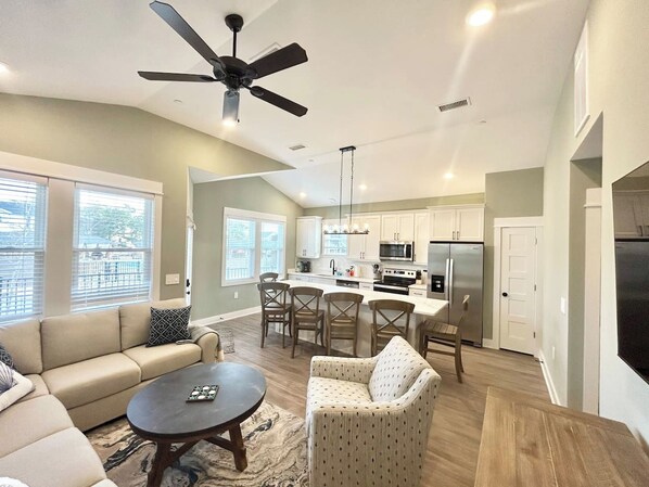 A modern living room with a sofa, armchair, and coffee table, a dining area with chairs, and a kitchen with appliances.
