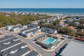 A residential area with a large swimming pool, multiple houses, and a beach in the background.