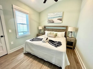 A bedroom with a bed, two bedside tables, a window with blinds, a painting on the wall, and a door.