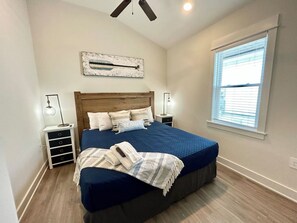 Une chambre à coucher comprenant un lit, des tables de chevet, un ventilateur de plafond, une fenêtre avec des stores et une œuvre d’art accrochée au mur.