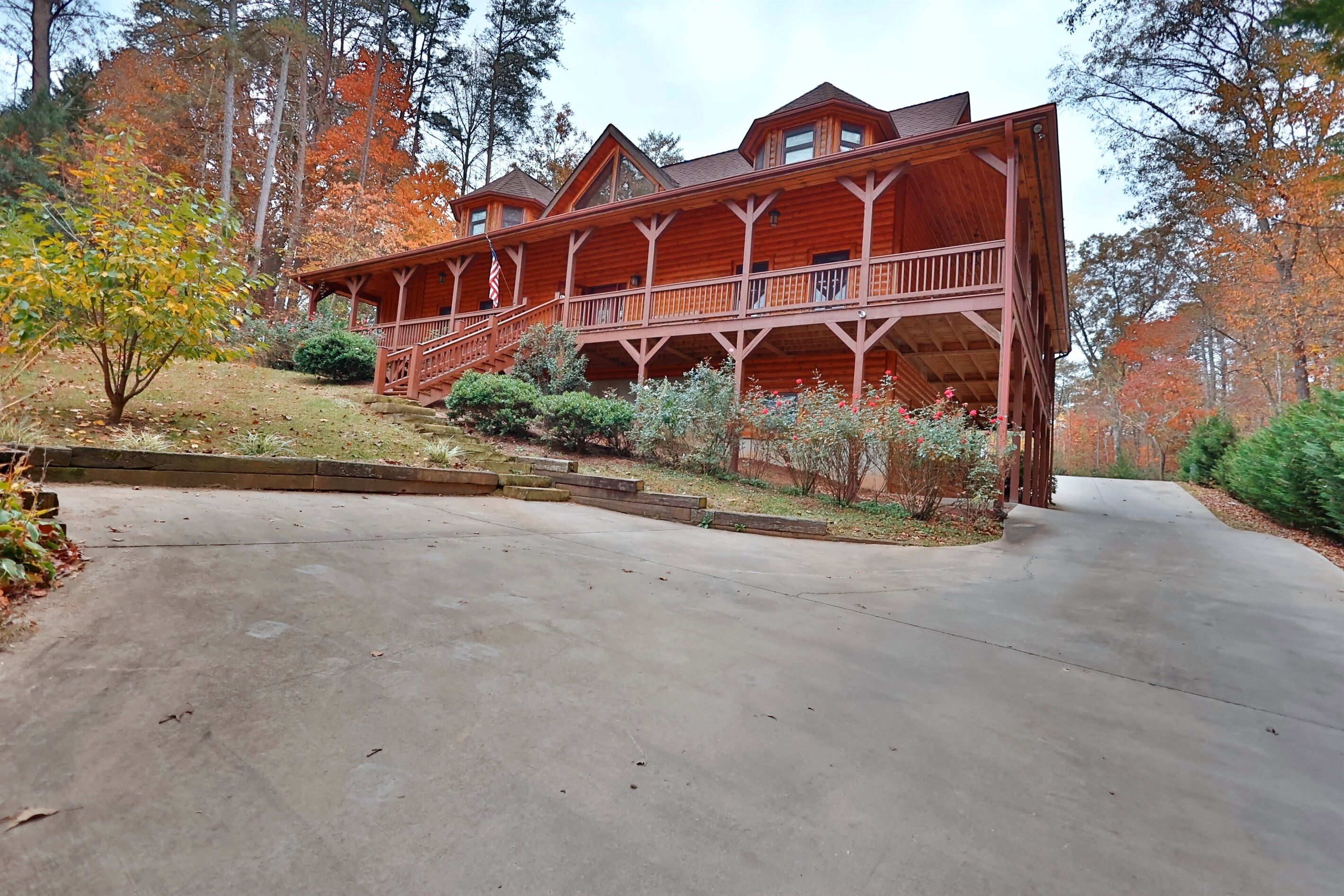 A large wooden cabin with a covered porch and multiple levels, surrounded by trees and a concrete driveway.