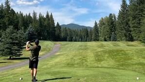 A golf course with a clear sky, a golfer in mid-swing, and a forested mountain in the background. - Private Mountain Retreat in the Woods! (Columbia Falls)
