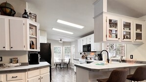 A modern kitchen with white cabinetry, a central island with bar stools, a microwave, and a large window providing natural light. - Private Mountain Retreat in the Woods! (Columbia Falls)