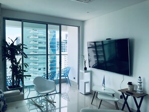 Un salon moderne avec une grande télévision, un balcon avec des chaises bleues et une plante en pot.