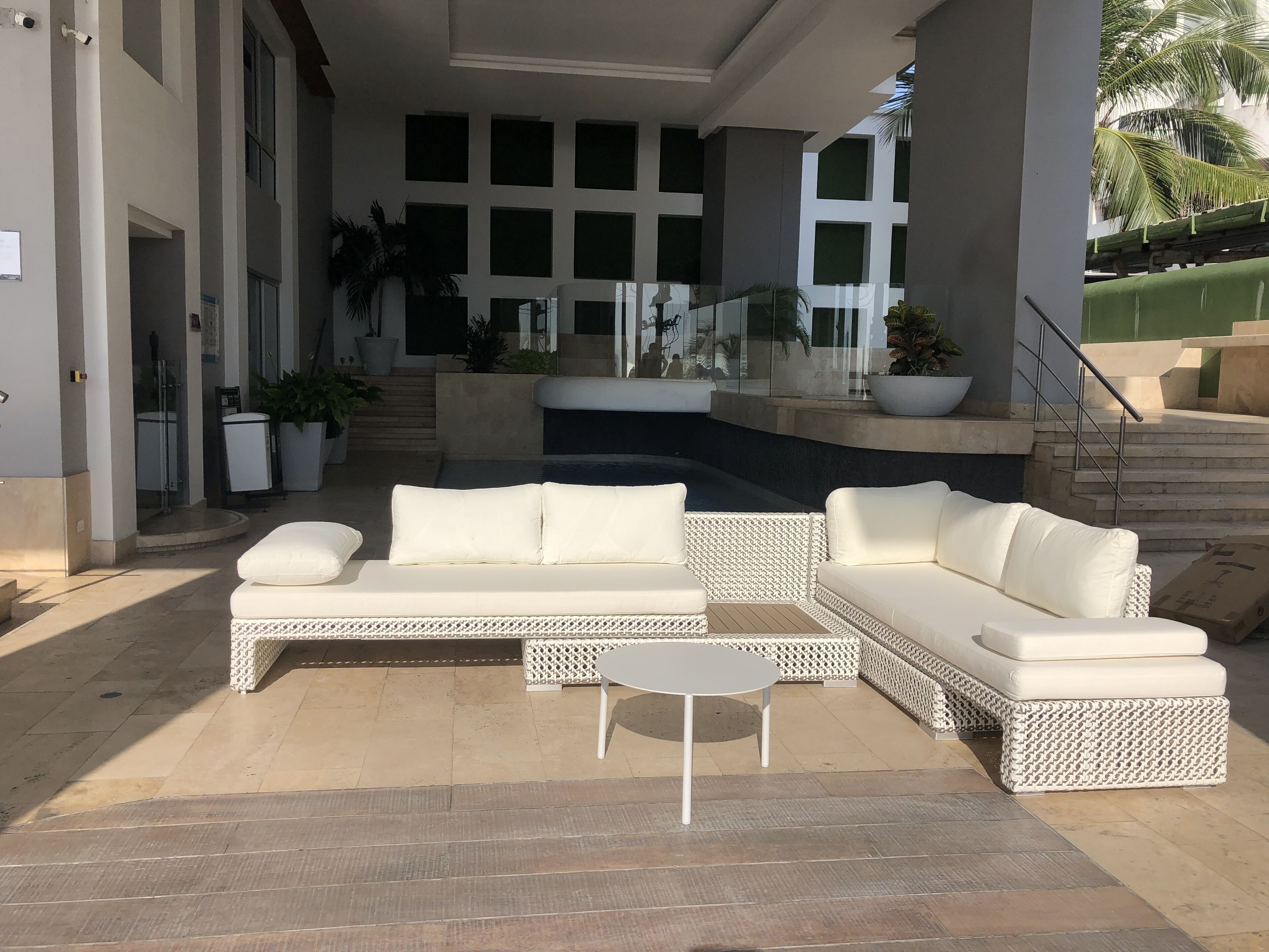 A modern outdoor lounge area with a white wicker sofa set, a small round table, and potted plants.