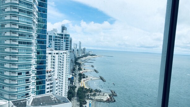 A coastal cityscape with modern high-rise buildings and a beach with umbrellas and people. - MURANO ELITE  GRADIMAR (Cartagena)