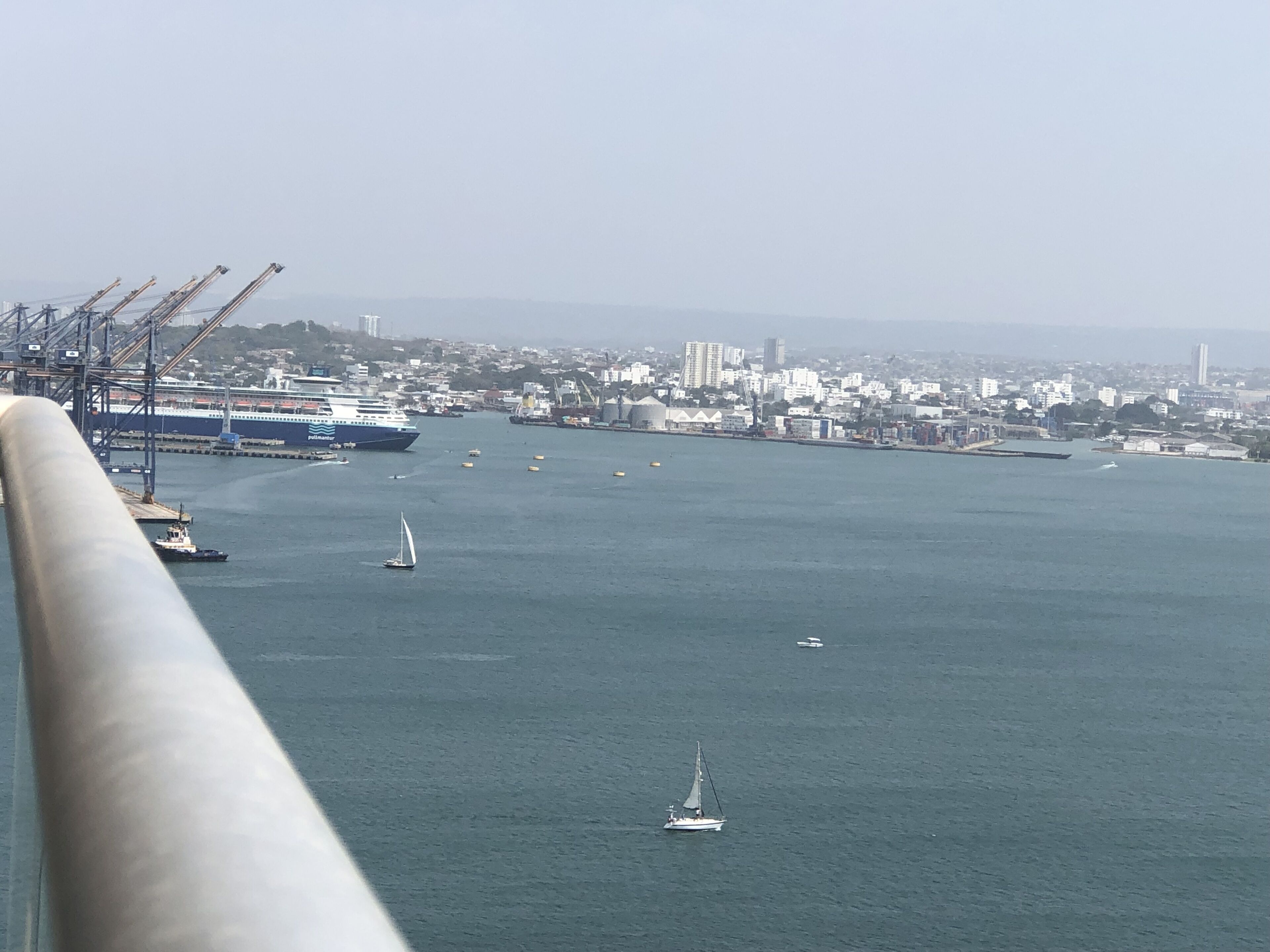 Um porto com navios, um veleiro e um horizonte urbano ao fundo.