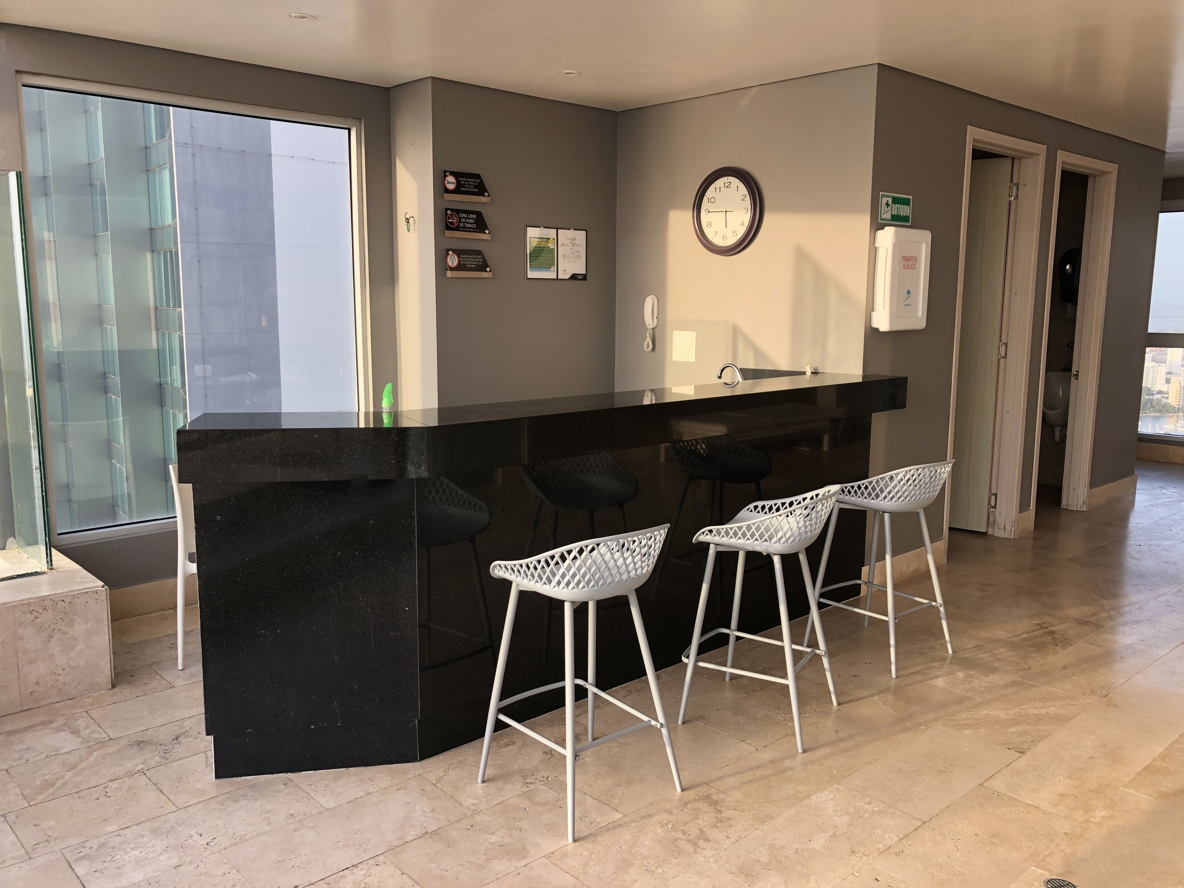 A modern kitchen with a black countertop, three white bar stools, a clock, and a window with a view of buildings.