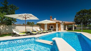 A swimming pool with lounge chairs and umbrellas, a covered patio, and a stone building with a tiled roof.