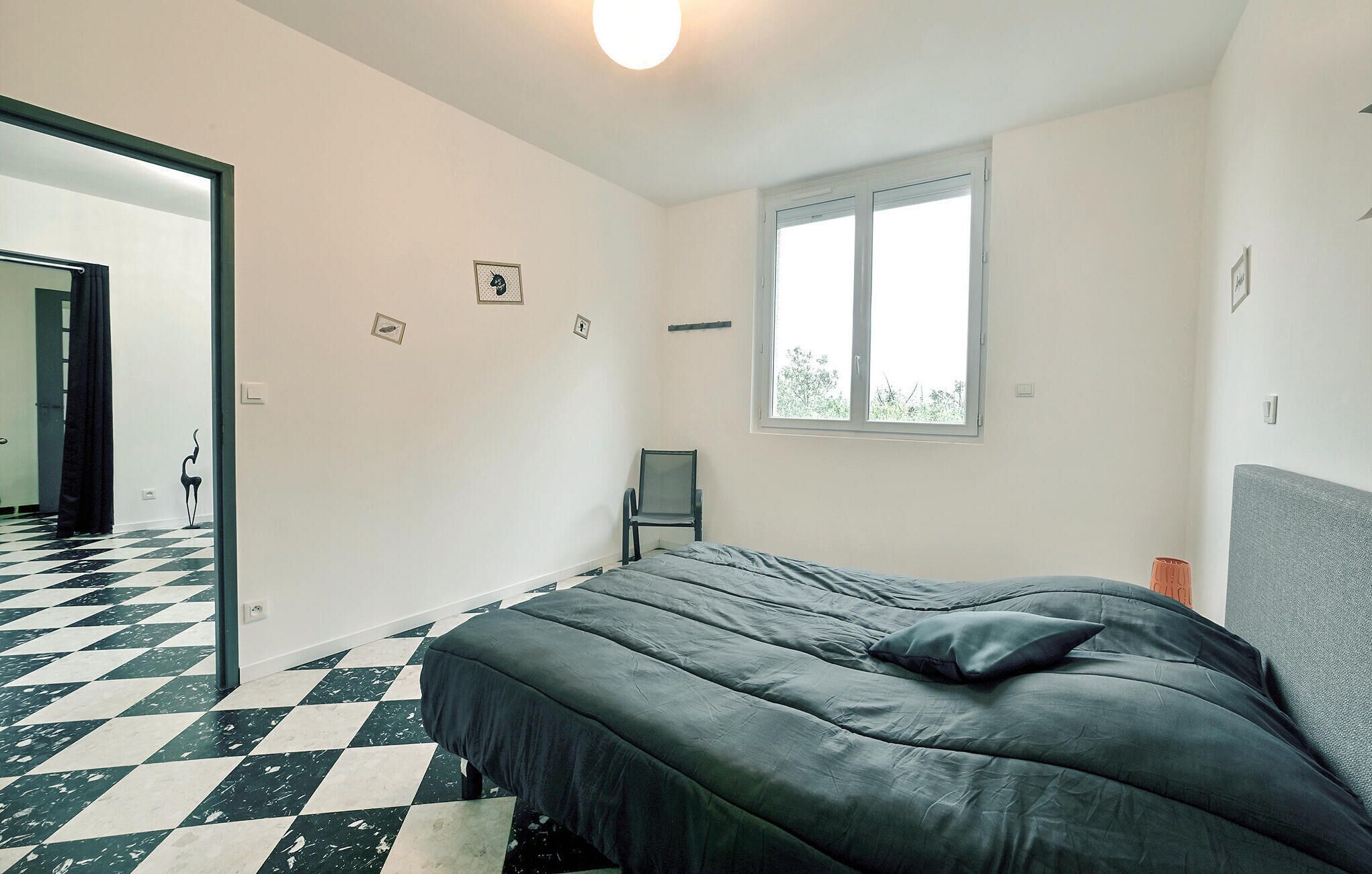 A bedroom with a checkered floor, a bed with a grey headboard, a window with a view of greenery, and a chair.