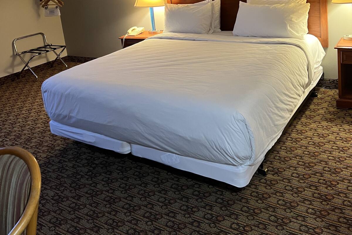 A hotel room with a large bed, two bedside lamps, a wooden headboard, a framed picture on the wall, a telephone on the nightstand, and a chair with a towel rack.