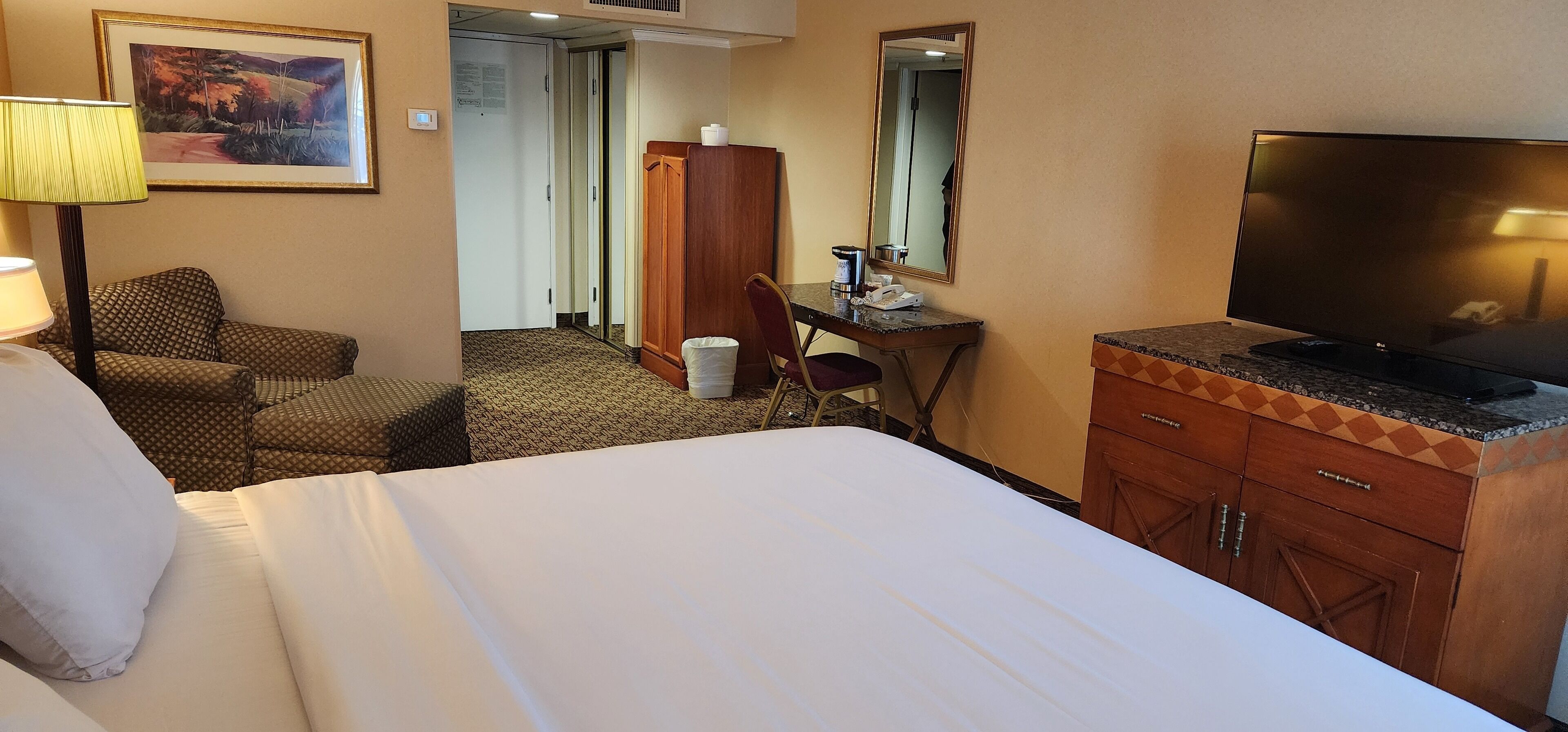 A hotel room with a bed, a desk with a chair, a television, a mirror, a lamp, and a painting on the wall.