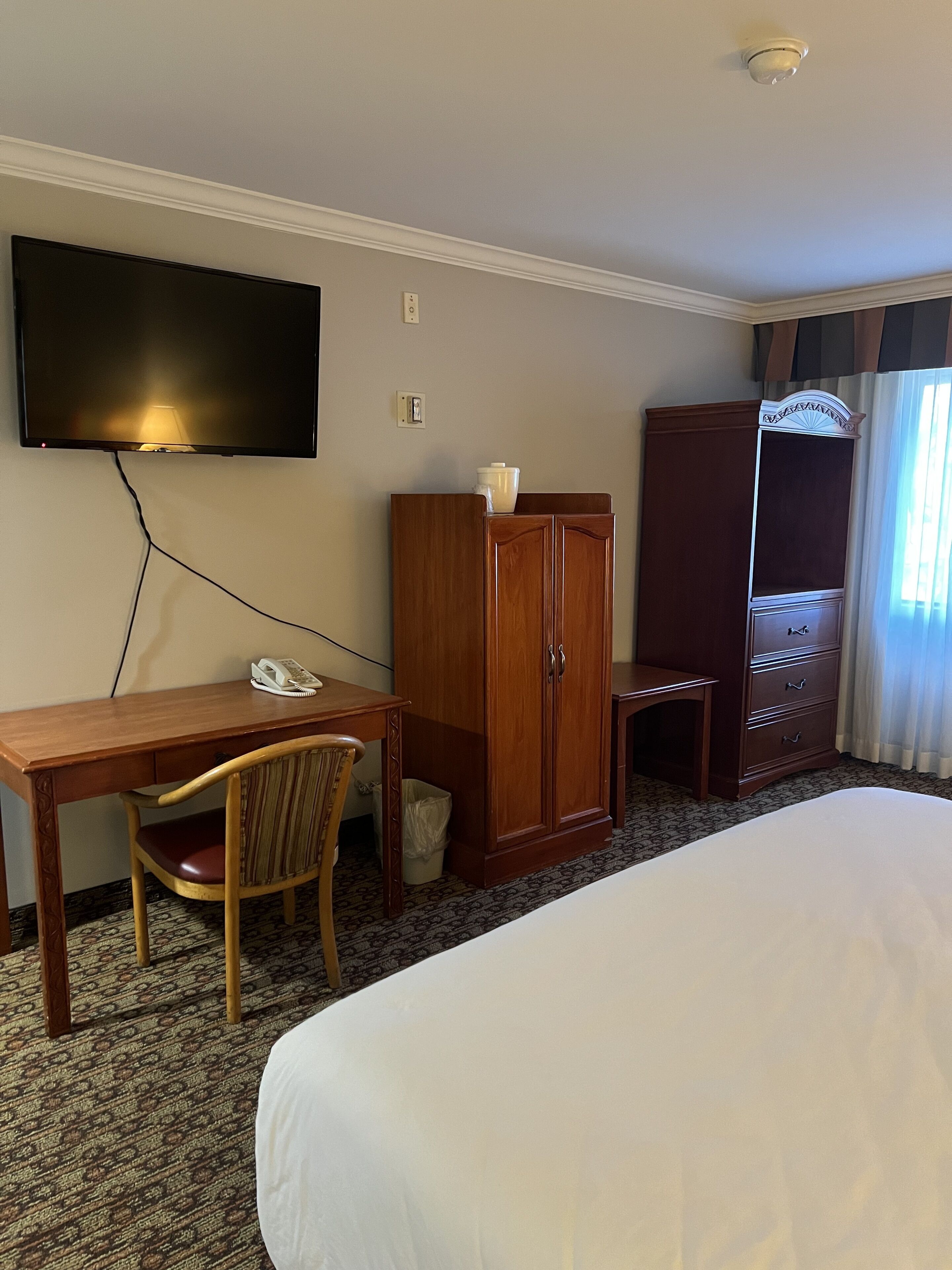 A hotel room with a bed, a desk with a telephone, a wooden wardrobe, and a television mounted on the wall.