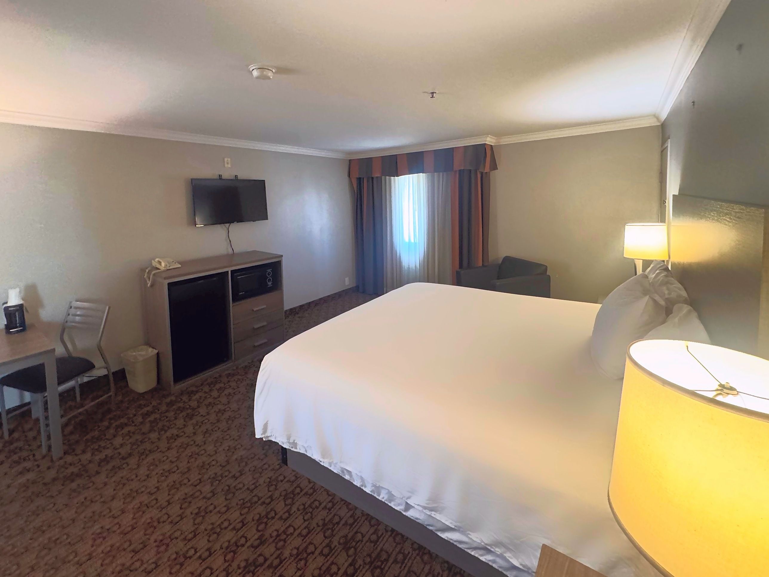 Habitación de hotel con cama, escritorio, televisor, silla, lámpara y ventana con cortinas.