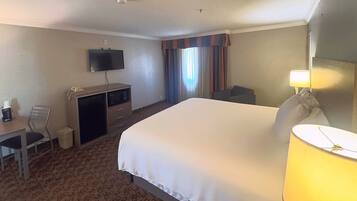 Habitación de hotel con cama, escritorio, televisor, silla, lámpara y ventana con cortinas.