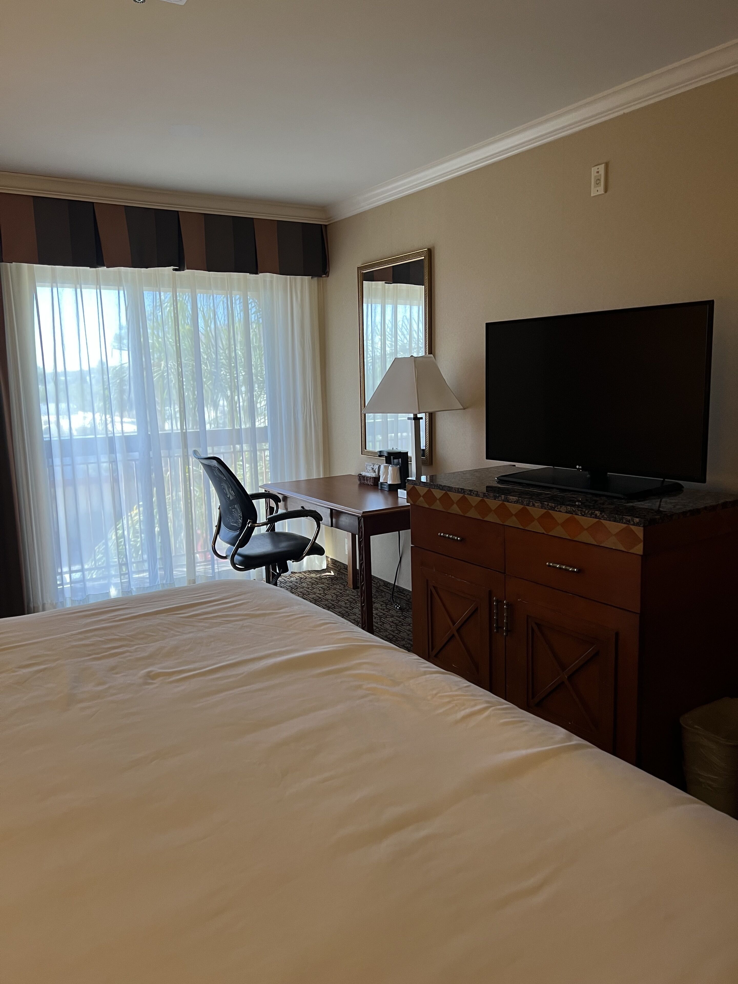 A hotel room with a bed, a desk with a lamp, a chair, a television, and a window with curtains.