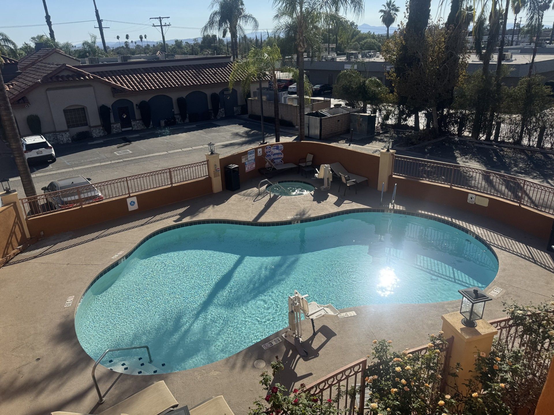 Una piscina con superficie de agua iluminada por el sol, rodeada por una valla baja y un área de descanso con sillas y una mesa.