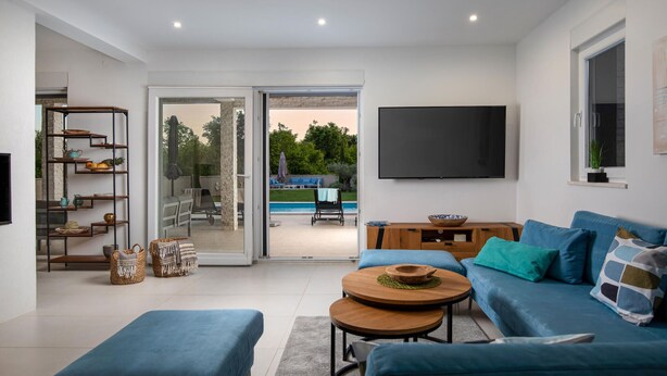A modern living room with a blue sofa, a coffee table, and a TV mounted on the wall. There is a sliding glass door leading to a pool area. - Nice home in Svetvincenat (Svetvincenat)