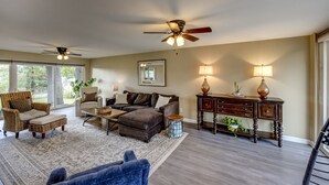 A spacious living room with a large sectional sofa, a coffee table, and a fireplace. There are two ceiling fans, a wooden cabinet, and a large window providing natural light. - Quiet Comfort: 5 BR & 2.5 BA, 6 TVs, Pet Friendly (Radcliff)