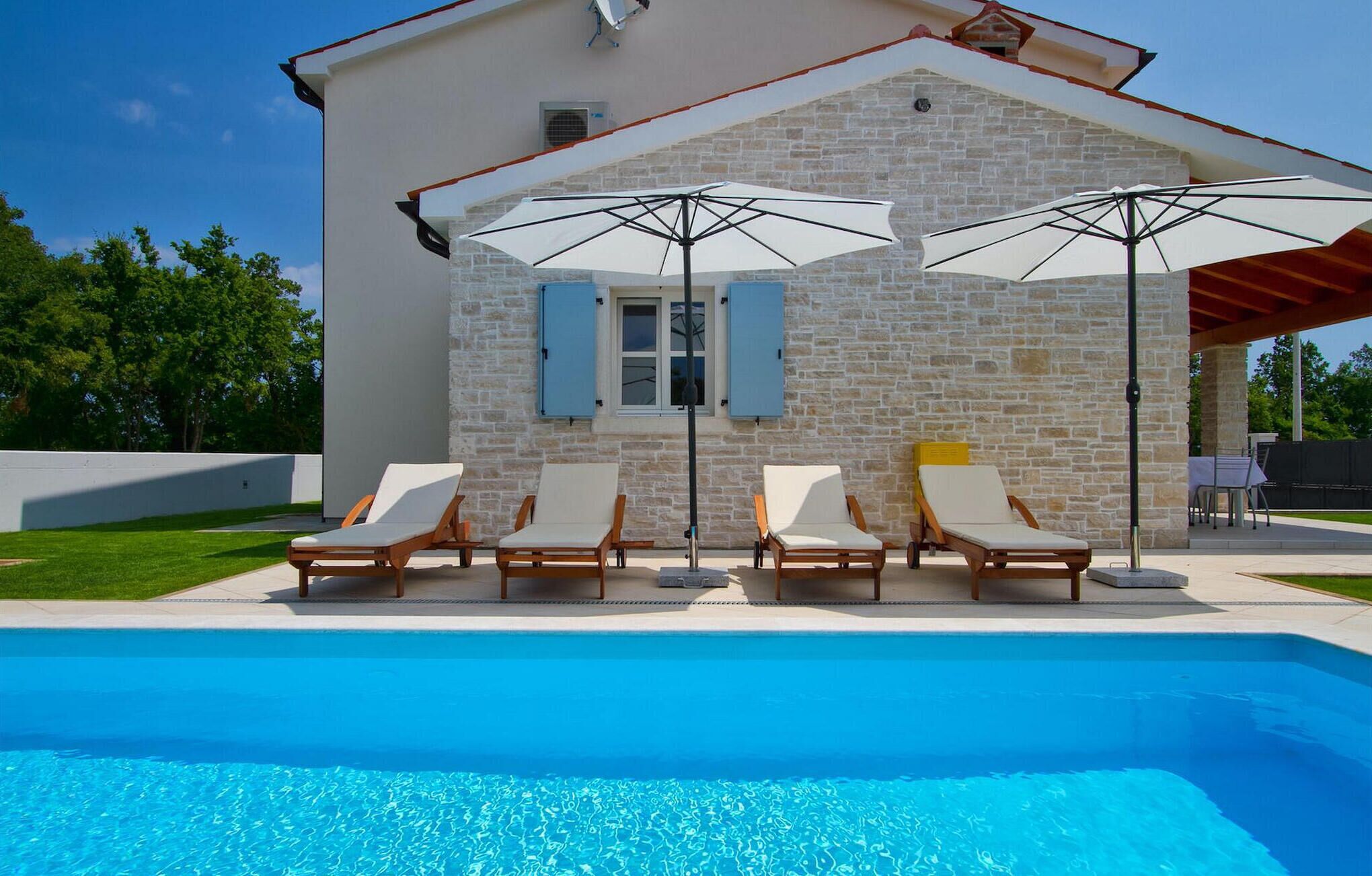 Un espace aménagé au bord de la piscine, avec des chaises longues et des parasols, un bâtiment en pierre et une piscine à l’eau cristalline.