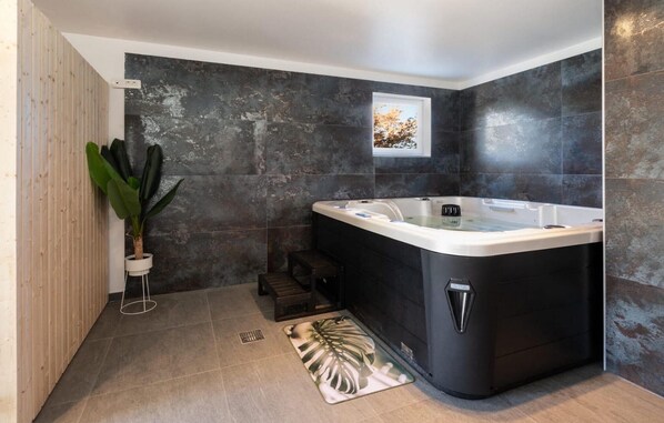 Un baño moderno con un jacuzzi negro grande, paredes de piedra oscura, un panel de madera en un lado y una ventana pequeña con vista a árboles.