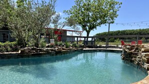 Una piscina cristalina con borde de piedra, rodeada de árboles y un edificio rojo al fondo.