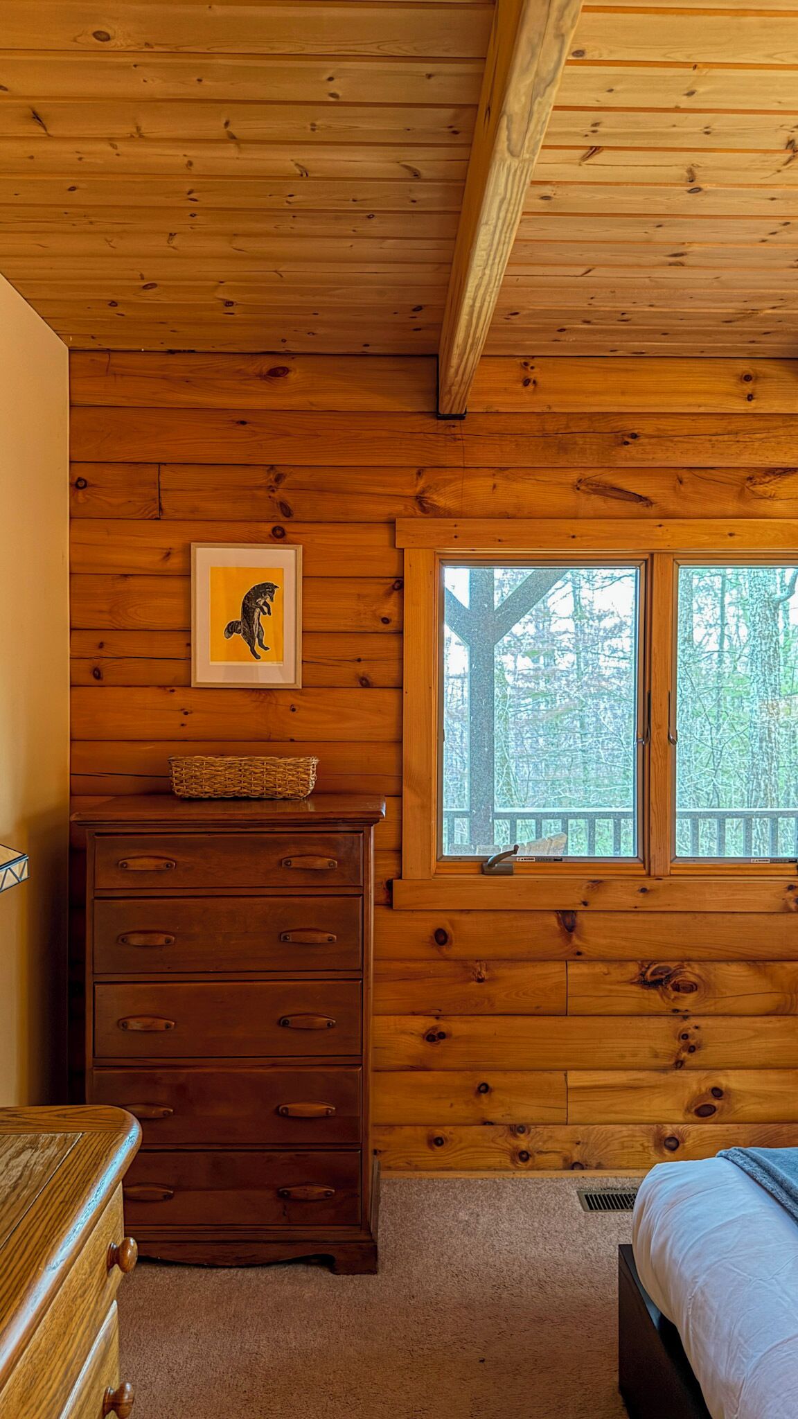 A wooden cabin room with a dresser, a framed picture, a window with a view of trees, and a bed.