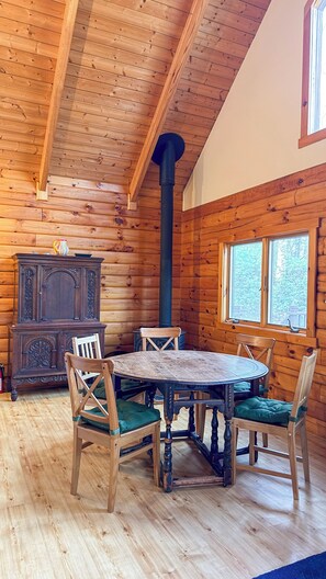 A wooden cabin interior with a round table and chairs, a tall chimney, and a window with a view of trees. - PERFECT ESCAPE For Total Privacy, Rustic Modern Cabin, 15 Pvt. Ac, WiFi, Heat/AC (Mt. Tremper)